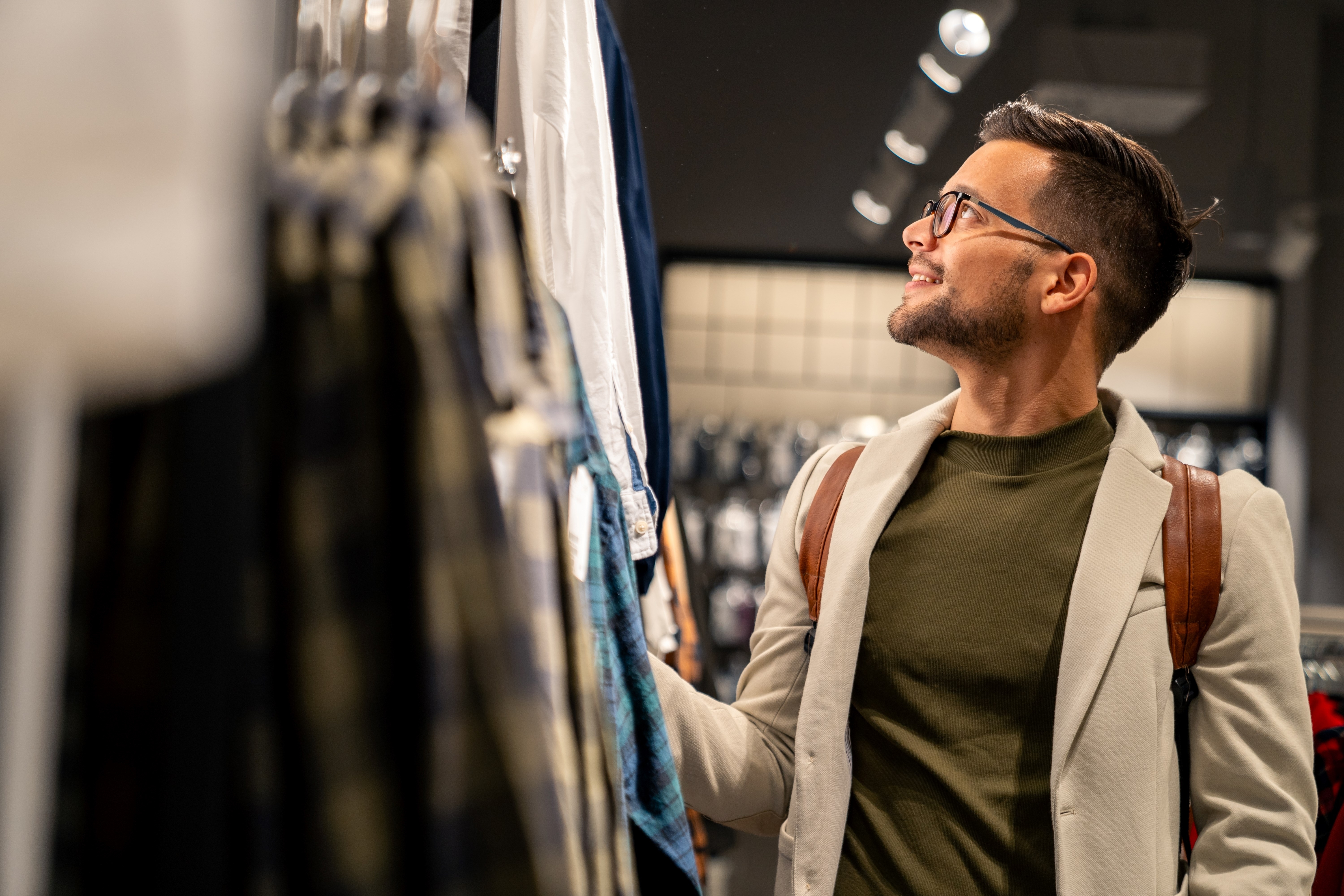 Caucasian male wearing glasses, olive shirt and beige blazer browses clothing selections in upscale retail store while carrying leather backpack.